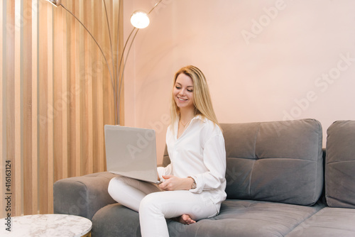 Beautiful young blonde smiling girl with laptop at home on the couch, remote work from home, isolation