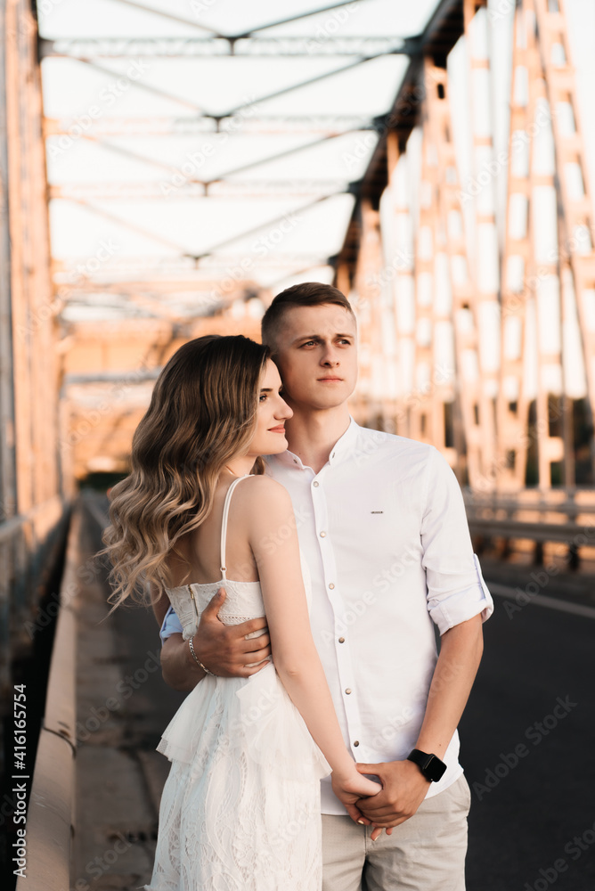 A beautiful young couple in love, a man and a woman, embrace, kiss on a large metal bridge at sunset.