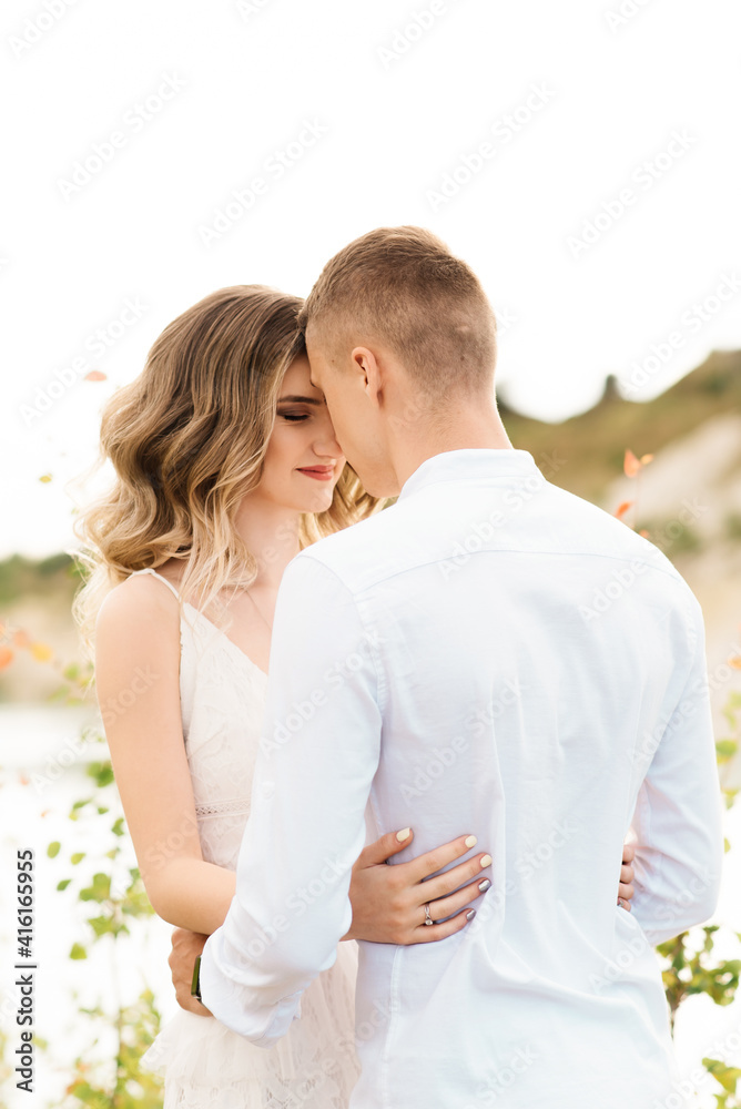 A beautiful young couple in love, a man and a woman embrace, kiss near a blue lake and sand at sunset. Vacation at the sea on the beach, sunset, morning, honeymoon, tropics, ocean, girl, relationship.