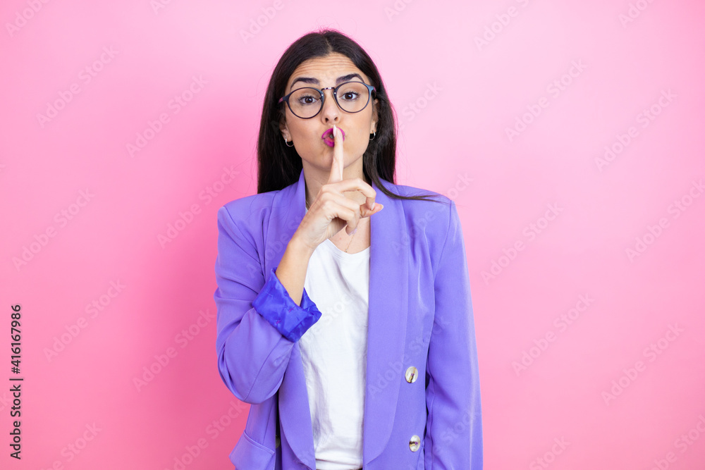 Young business woman wearing purple jacket over pink background asking to be quiet with finger on lips. Silence and secret concept. Young business woman wearing purple jacket over pink background asking to be quiet with finger on lips. Silence and secret concept.