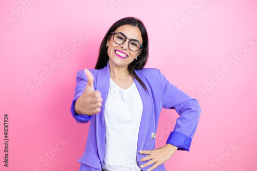 Young business woman wearing purple jacket over pink background smiling friendly offering handshake as greeting and welcoming