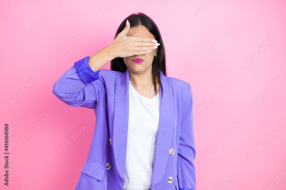 Young business woman wearing purple jacket over pink background serious and covering her eyes with her hand Young business woman wearing purple jacket over pink background serious and covering her eyes with her hand