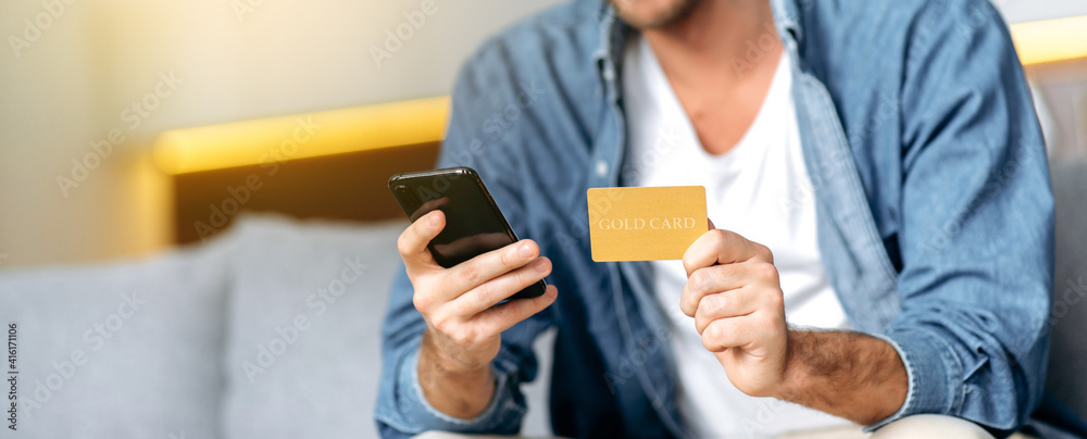 Panoramic photo of a credit card and smartphone in male hands. Caucasian young man, is using mobile phone and credit card for online shopping while sitting at home on sofa. Credit card and phone in Panoramic photo of a credit card and smartphone in male hands. Caucasian young man, is using mobile phone and credit card for online shopping while sitting at home on sofa. Credit card and phone in