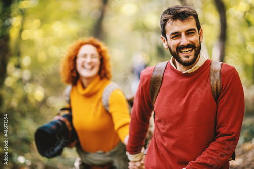 Young smiling happy couple holding hands and walking in nature on a beautiful autumn day. Selective focus on man. Couple is holding picnic equipment and having backpacks on backs.