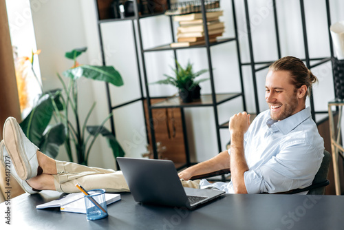 Smiling attractive young man, freelancer or manager, is relaxing at workplace with legs thrown on table. Modern guy uses laptop, browses the Internet, looks for ideas for a project, while sitting at © Kateryna