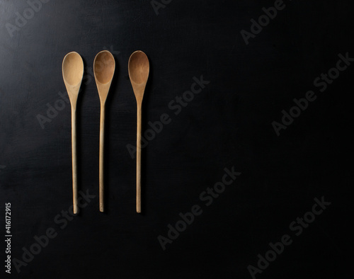 Three wooden spoons in different shades of brown used in the kitchen to cook, season and taste new flavors. On a gray table with lighting that transcends horizontally. © studioneeby