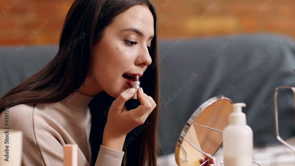 Young self-isolated woman applying new lipstick on lips, looking at table mirror, sitting at home