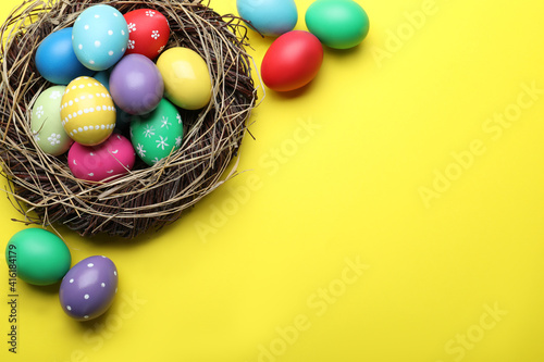 Bright painted eggs in nest on yellow background, flat lay with space for text. Happy Easter
