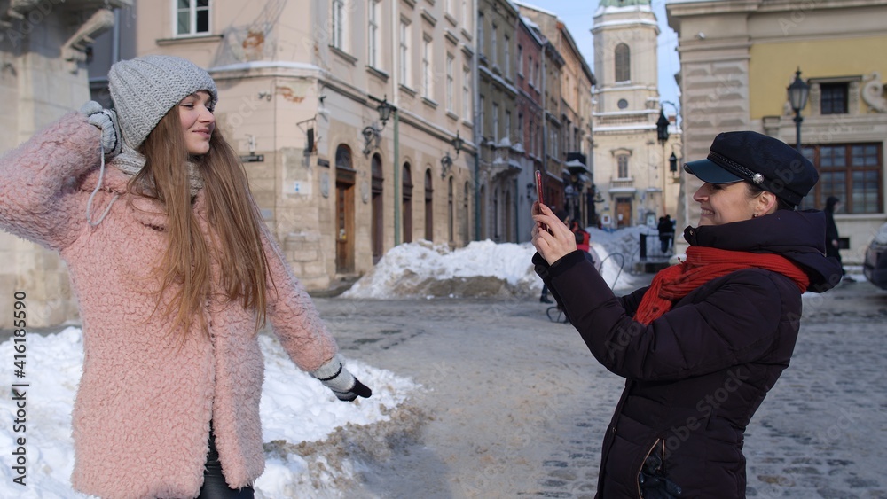 Fototapeta premium Two young smiling women tourists bloggers taking photos portrait on mobile phone while standing on city street. Happy lesbian LGBT couple during winter holidays traveling vacation making online vlog
