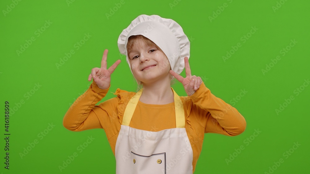Little child girl dressed in apron and hat like chef cook showing victory sign, hoping for success and win, doing peace gesture and smiling with kind optimistic expression on chroma key background Little child girl dressed in apron and hat like chef cook showing victory sign, hoping for success and win, doing peace gesture and smiling with kind optimistic expression on chroma key background