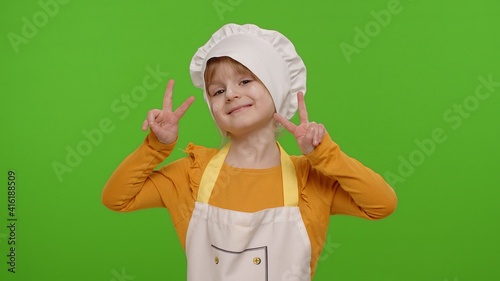 Little child girl dressed in apron and hat like chef cook showing victory sign, hoping for success and win, doing peace gesture and smiling with kind optimistic expression on chroma key background
