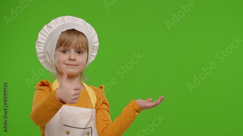 Funny preschooler child girl dressed cook chef baker in apron and hat pointing at right on blank space on chroma key background. Place for your advertisement logo. Cooking school, children education photo