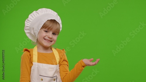 Child girl dressed in apron and hat like chef cook pointing at right on blank space. Place for your advertisement logo. Chroma key background. Nutrition, family cooking school, children education
