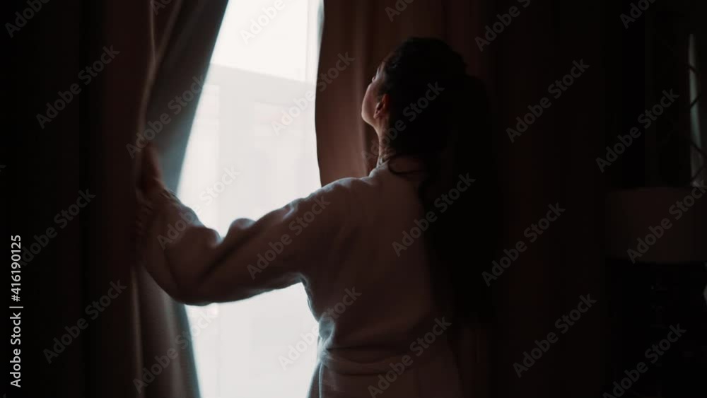 A young attractive brunette walks to the window and opens the curtains. Fresh lazy lady wake up after healthy sleep at home. Enjoys beautiful daylight, beautiful mornings and weekends A young attractive brunette walks to the window and opens the curtains. Fresh lazy lady wake up after healthy sleep at home. Enjoys beautiful daylight, beautiful mornings and weekends