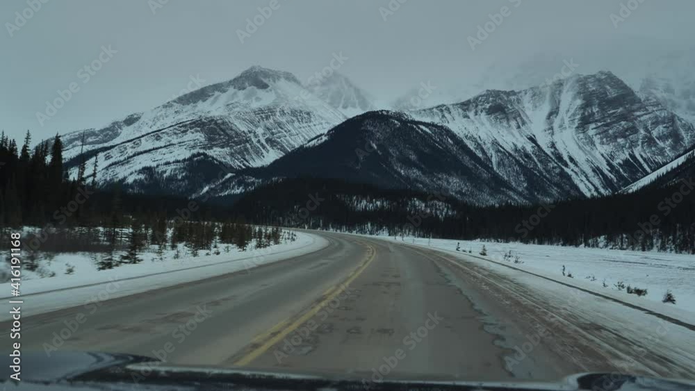 Driver perspective view of a snowy road with a car driving through the forest in the Canadian Rocky Mountains in Jasper and Banff National Park, Alberta, Canada Driver perspective view of a snowy road with a car driving through the forest in the Canadian Rocky Mountains in Jasper and Banff National Park, Alberta, Canada