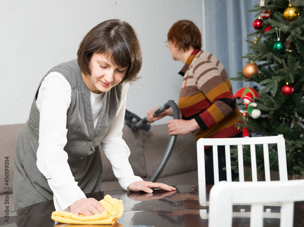 Attractive woman wiping table surface while her elderly mother vacuum cleaning sofa in preparation for celebration of New Year..