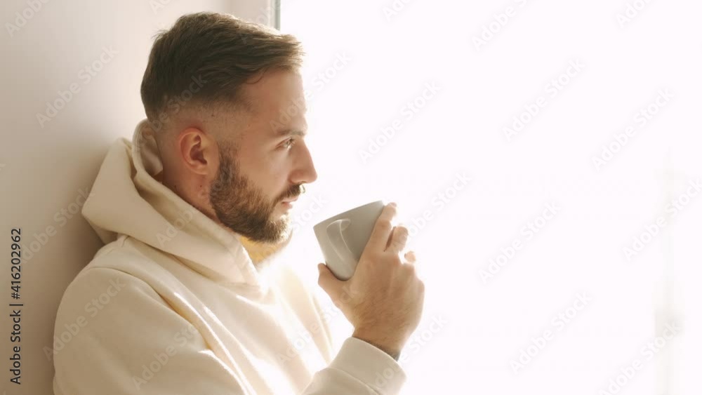 Young man resting on chair with mobile phone during his leisure time. Man with cup of tea relaxing near window at home Young man resting on chair with mobile phone during his leisure time. Man with cup of tea relaxing near window at home