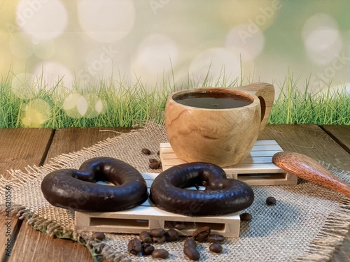 An oak table with visible grains is standing on a wood cup with black coffee. On a jute napkin, coffee beans are scattered and a gingerbread bitten heart lies on the pallet 