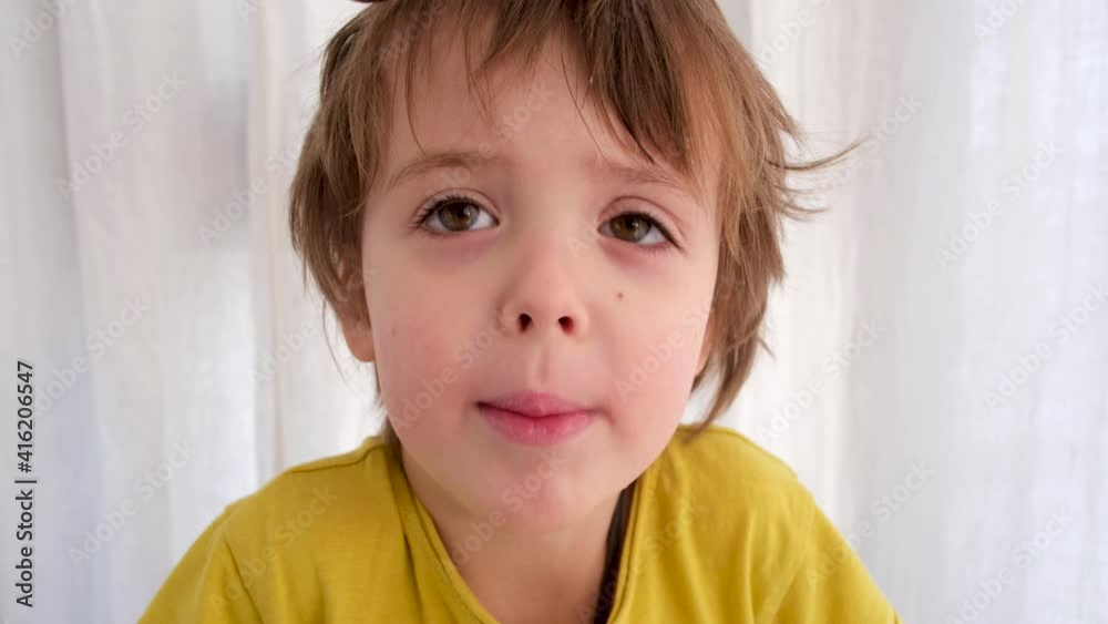 Adorable happy little boy wearing yellow t-shirt shrugs posing for camera against white window curtain in light room closeup Adorable happy little boy wearing yellow t-shirt shrugs posing for camera against white window curtain in light room closeup