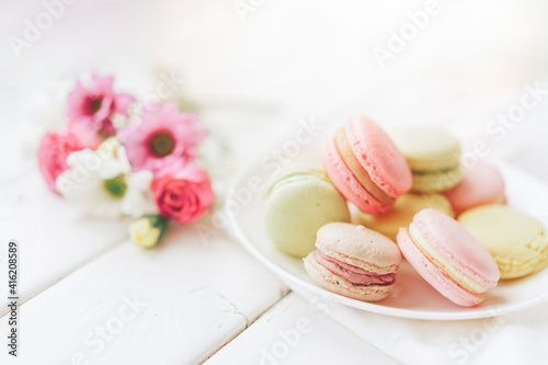 A full plate of macaroons lie in a plate of different colors, next to the plate is a small bouquet of flowers against a white background