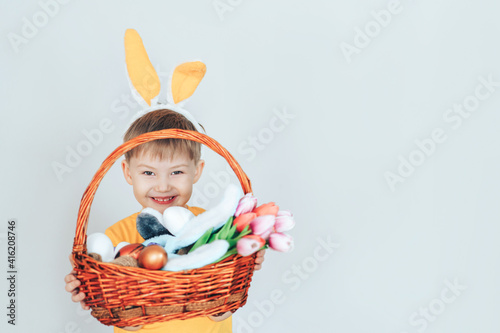 A little boy in a rabbit costume holding a large basket filled with eggs and tulips against a white background