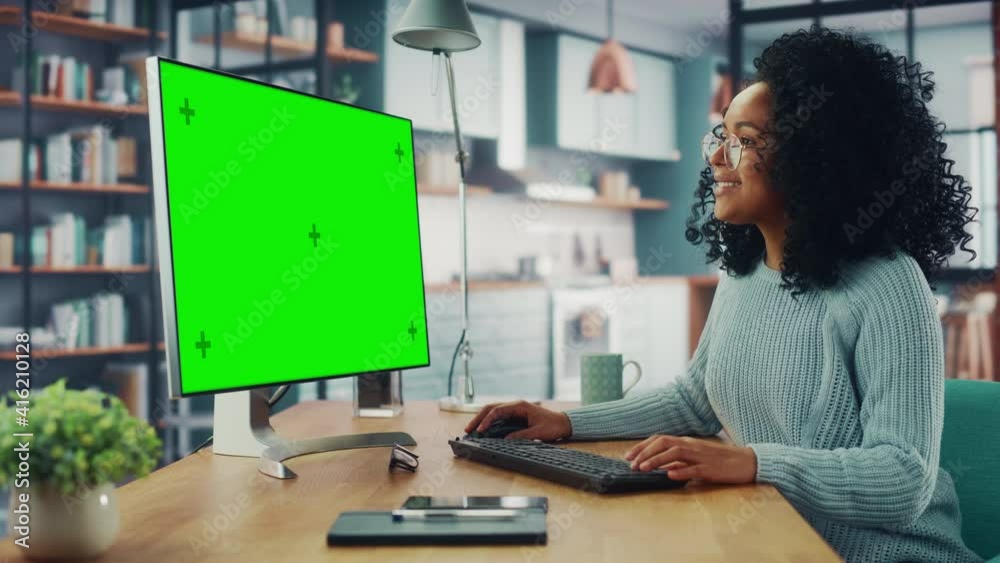 Latina Female Specialist Working on Desktop Computer with Green Screen Mock Up Display at Home Living Room while Sitting at a Table. Freelancer Female Chatting Over the Internet on Social Networks.