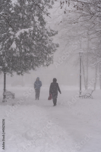 City park in winter with heavy snowfall. Passers people walks down the street in the snow storm in the city.