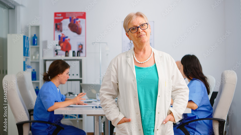 Obraz premium Portrait of senior medic looking at camera smiling standing in medical conference meeting office. Team of specialist doctors working in background talking about symptoms of disease in clinic room