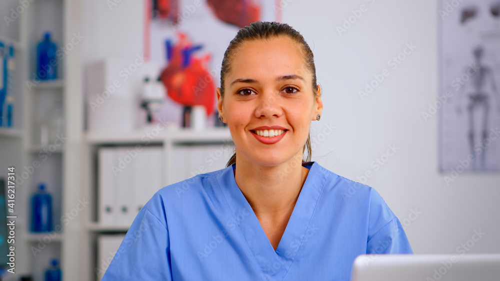 Portrait of nurse looking at camera smiling after typing on laptop sitting in hospital office wearing blue uniform. Hospital healthcare worker, medicine clinical assistant health consultant therapist Portrait of nurse looking at camera smiling after typing on laptop sitting in hospital office wearing blue uniform. Hospital healthcare worker, medicine clinical assistant health consultant therapist