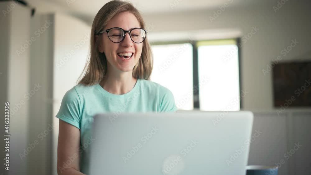 Close-up shot of a beautiful young adult cheerful woman in glasses taking a video call on a laptop indoors.