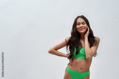 Studio shot of beautiful young woman wearing green underwear smiling with eyes closed at camera, posing isolated over light background © Svitlana