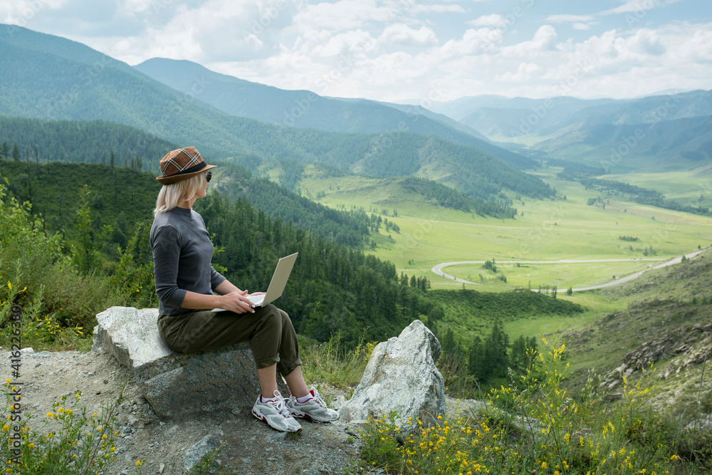Fototapeta premium Beautiful woman hipster traveler freelancer with laptop on the top of the mountain. Remote work concept.