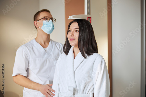 Serious bespectacled physician in a face mask applying a stadiometer photo