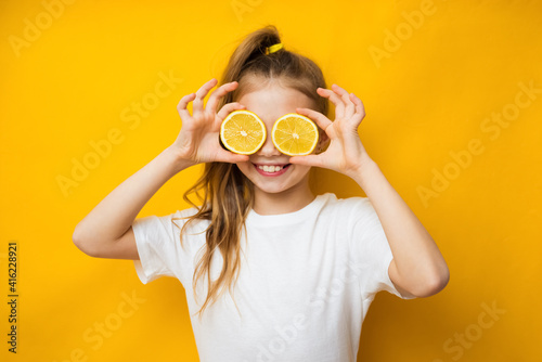 Little smiling cute blond girl in yellow t-shirt holding halves of fresh sour lemon fruit near eyes and showing tongue over yellow background. Healthy lifestyle and clean eating concept