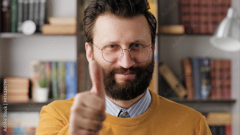 Naklejka premium Man thumb up. Positive smiling bearded man in glasses in office or apartment room looking at camera and shows his thumb up. Close-up