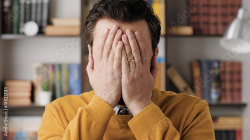 FACEPALM. Astonished bearded man in glasses in office or apartment room looks at camera and covers his face with his hands expressing his bewilderment. Close-up