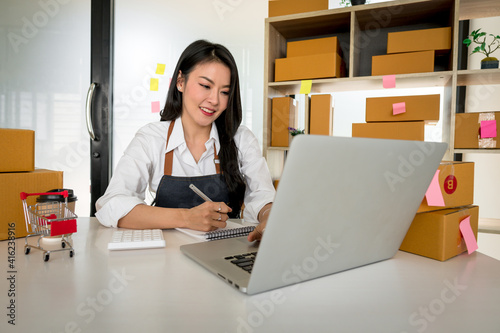 Portrait, entrepreneur, young Asian woman, freelance business, sme business, online shopping, working on laptop computer with parcel box on home office table, online business and delivery concept.