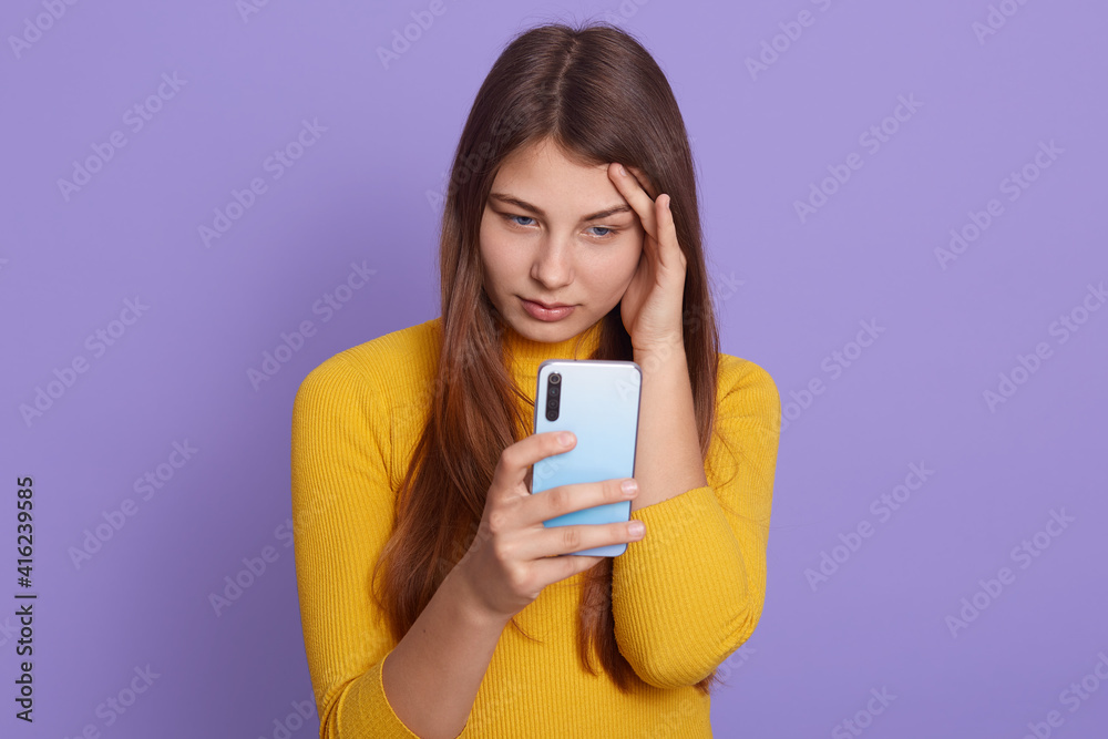 Pretty European woman ponders on how to answer question, read bad news, has problems, touch her temple with fingers, thinks deeply about something, uses mobile phone, stands indoor against lilac wall. Pretty European woman ponders on how to answer question, read bad news, has problems, touch her temple with fingers, thinks deeply about something, uses mobile phone, stands indoor against lilac wall.