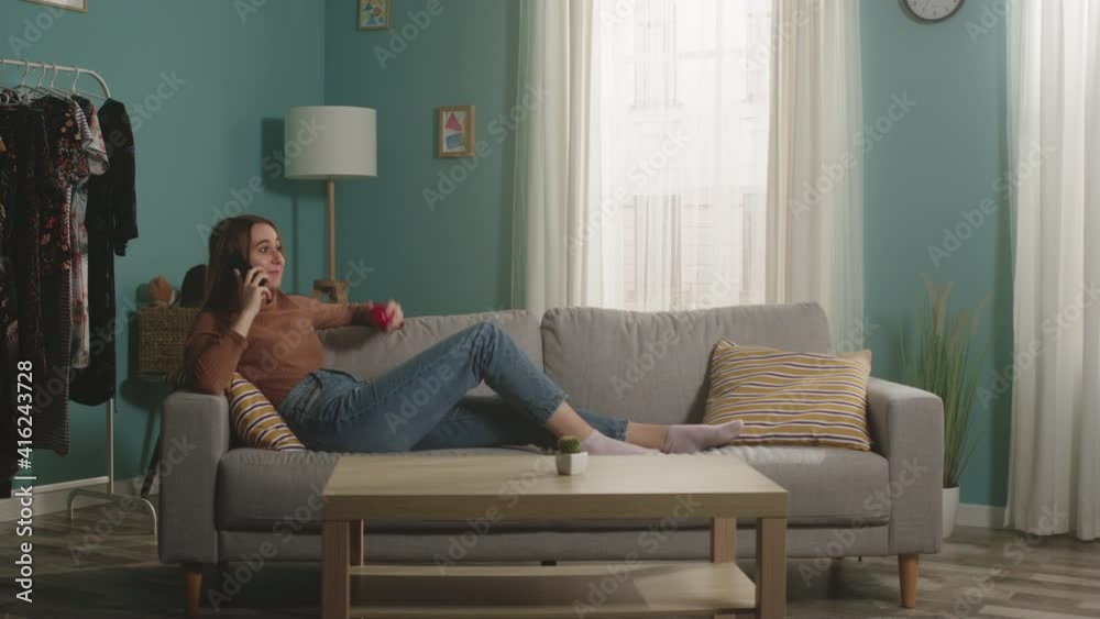 Beautiful woman in blue jeans and a brown sweater lies on a beige sofa on the background of the window, holding a red apple and talking on the phone. General plan. Beautiful woman in blue jeans and a brown sweater lies on a beige sofa on the background of the window, holding a red apple and talking on the phone. General plan.