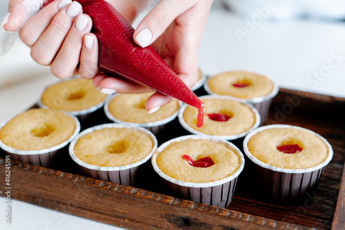 the hands of a female pastry chef hold a cooking bag and fill the red berry filling cupcakes in a wooden tray.Food for breakfast. Freshly baked cupcakes for dessert. Foodies and cuisine.