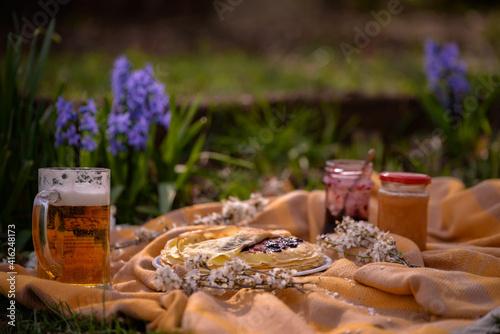 picnic in the spring season on the green grass in the flower garden. a pint of beer placed on the blanket next to a plate of pancakes with jam