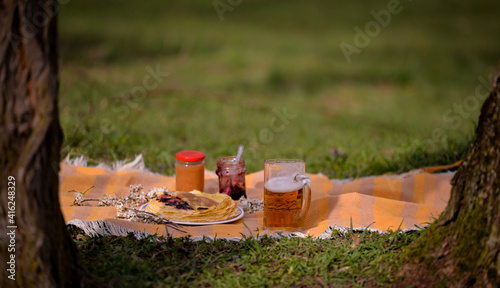 picnic in the spring season on the green grass in the flower garden. a pint of beer placed on the blanket next to a plate of pancakes with jam