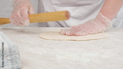 The chef's hands in gloves roll out the dough with a wooden dough roller. Tight dough for dumplings, ravioli.
