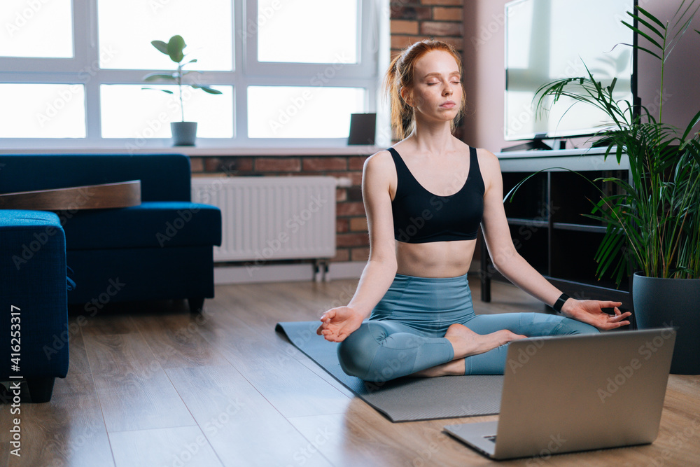Calm redhead young woman with closed eyes meditating at home sitting in lotus position on yoga mat near laptop computer. Peaceful red-haired lady holding hands on knee in Om position. Calm redhead young woman with closed eyes meditating at home sitting in lotus position on yoga mat near laptop computer. Peaceful red-haired lady holding hands on knee in Om position.