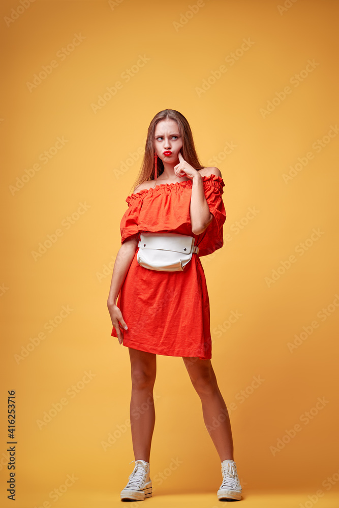 Summer urban fashion. Fun and colorful. Young pretty happy woman in orange dress posing against yellow background. Summer urban fashion. Fun and colorful. Young pretty happy woman in orange dress posing against yellow background.