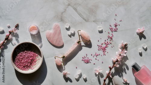 Crystal rose quartz facial roller and Gua sha stone for beauty facial massage therapy, Flat lay on marble table with magnolia flowers. Long shadows and candle. Essential oil bottle. Panoramic image.
