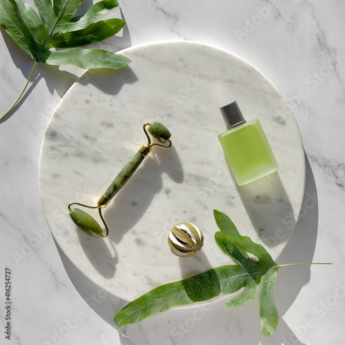 Jade face roller for facial massage therapy, Essential oil glass bottle. Top view on round marble podium on matching stone table with exotic leaves of ferns. Simple arrangement, sunlight, long shadows