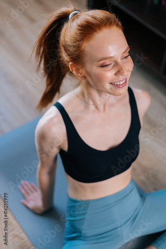 High-angle view of cheerful redhead young woman wearing sportswear sitting in floor on yoga mat, looking away. Concept of sports training red-haired lady during quarantine.