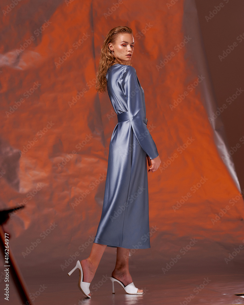 Elegant blonde woman with fashionable make-up and long wet hair in blue silk elegant dress standing and posing against red orange metallic background