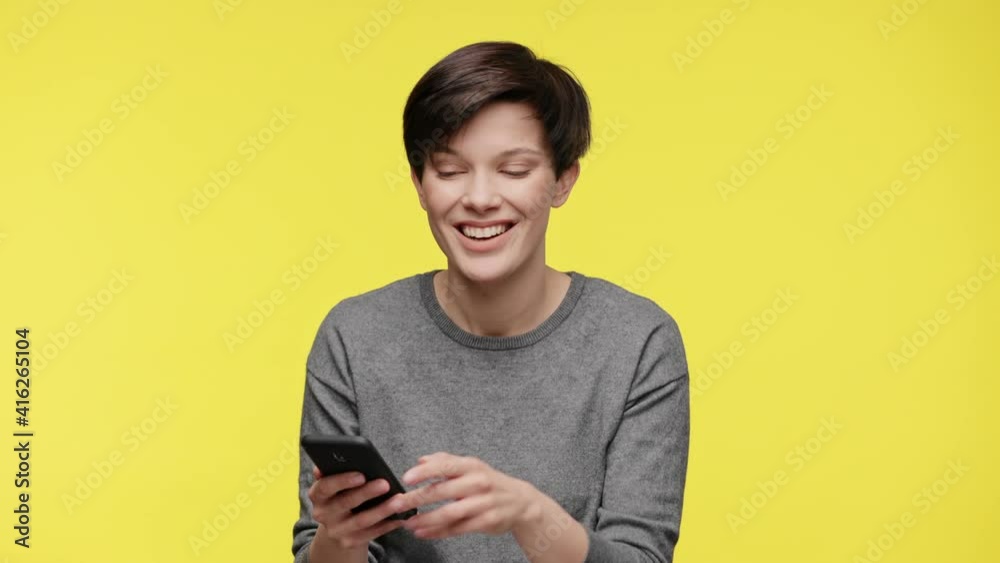 Color of the year 2021. Ultimate Gray and illuminating. Surprised young beautiful woman in gray sweatshirt using smartphone, get shocking and unbelievable news, isolated on yellow background. Color of the year 2021. Ultimate Gray and illuminating. Surprised young beautiful woman in gray sweatshirt using smartphone, get shocking and unbelievable news, isolated on yellow background.